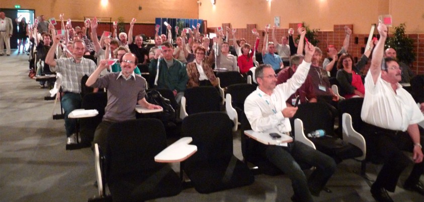 Une assemblée de personnes assises dans une salle brandissent une vague de carrés rouges.