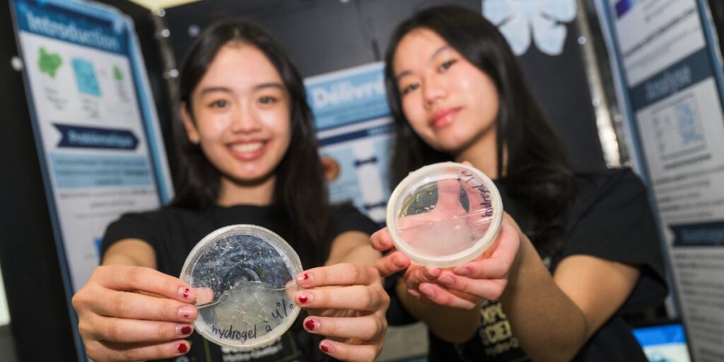 Devant une affiche scientifiques, deux jeunes filles aux visages flous tiennent devant elles des boîtes de Pétri contenant des hydrogels.